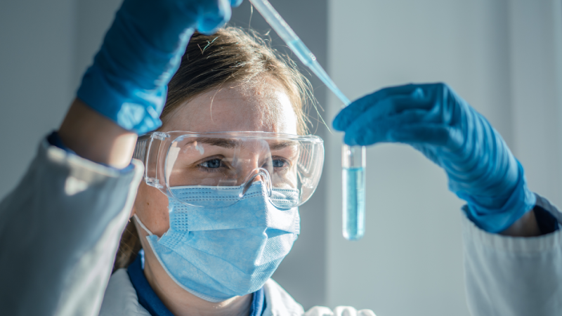 Female scientist wearing safety equipment carefully transfers liquid with pipette in laboratory setting. Professional researcher performs chemical analysis in bright, sterile environment