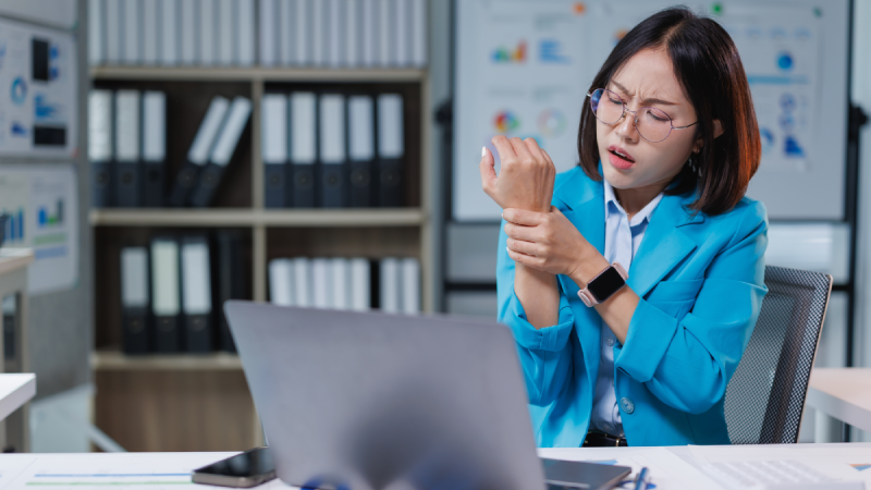 Businesswoman experiencing wrist pain while working long hours on a laptop in an office, indicating symptoms of carpal tunnel syndrome or a work-related injury