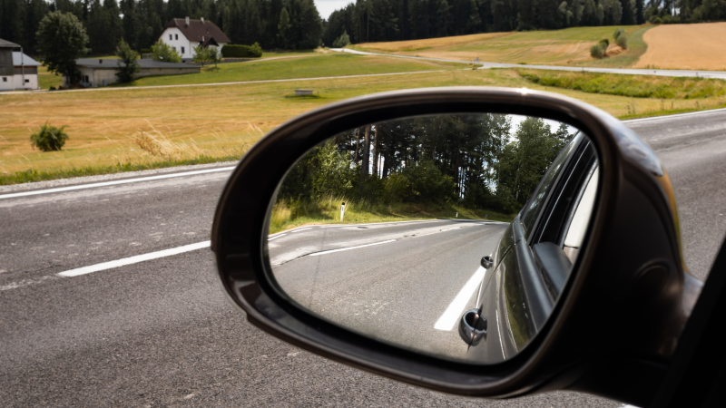 View into the car side mirror on the left side. The street in the rural area is empty. The blind spot is not visible in the rearview mirror
