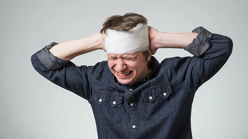 Traumatized young guy with bandage on his head putting hands on wound.