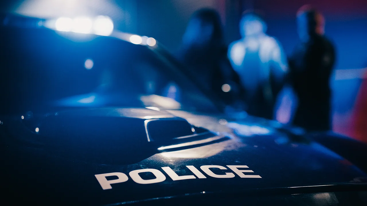 the hood of a police cruiser with flashing blue emergency lights is in sharp focus, while police officers and a suspect stand blurred in the background at night.